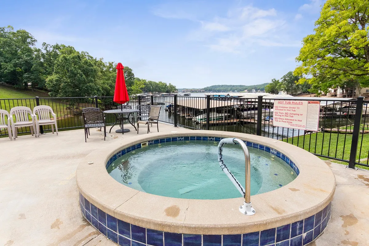 A picturesque hot tub scene overlooks a beautiful waterfront view, with a red umbrella providing shade and an outdoor seating area nearby. The setting invites relaxation and socializing, whether it's soaking in the hot tub under the stars or enjoying the surrounding natural scenery.