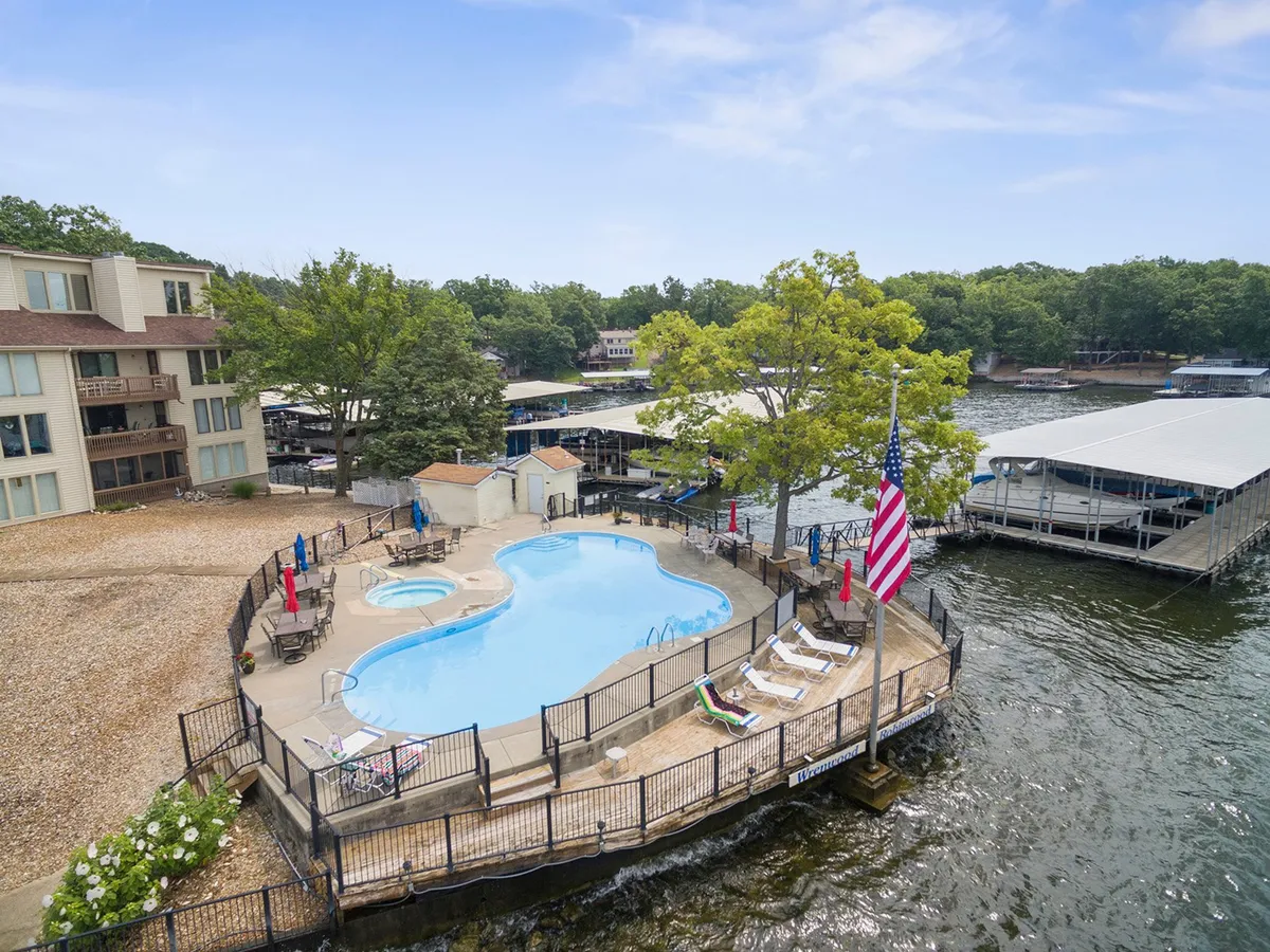 A vibrant scene of a lakefront swimming pool flanked by a dock and lounge chairs, with a flag waving in the breeze and mature trees in the background. The mood is leisurely, suggesting activities like swimming, sunbathing, boating, and gathering with friends as the sun sets over the water.