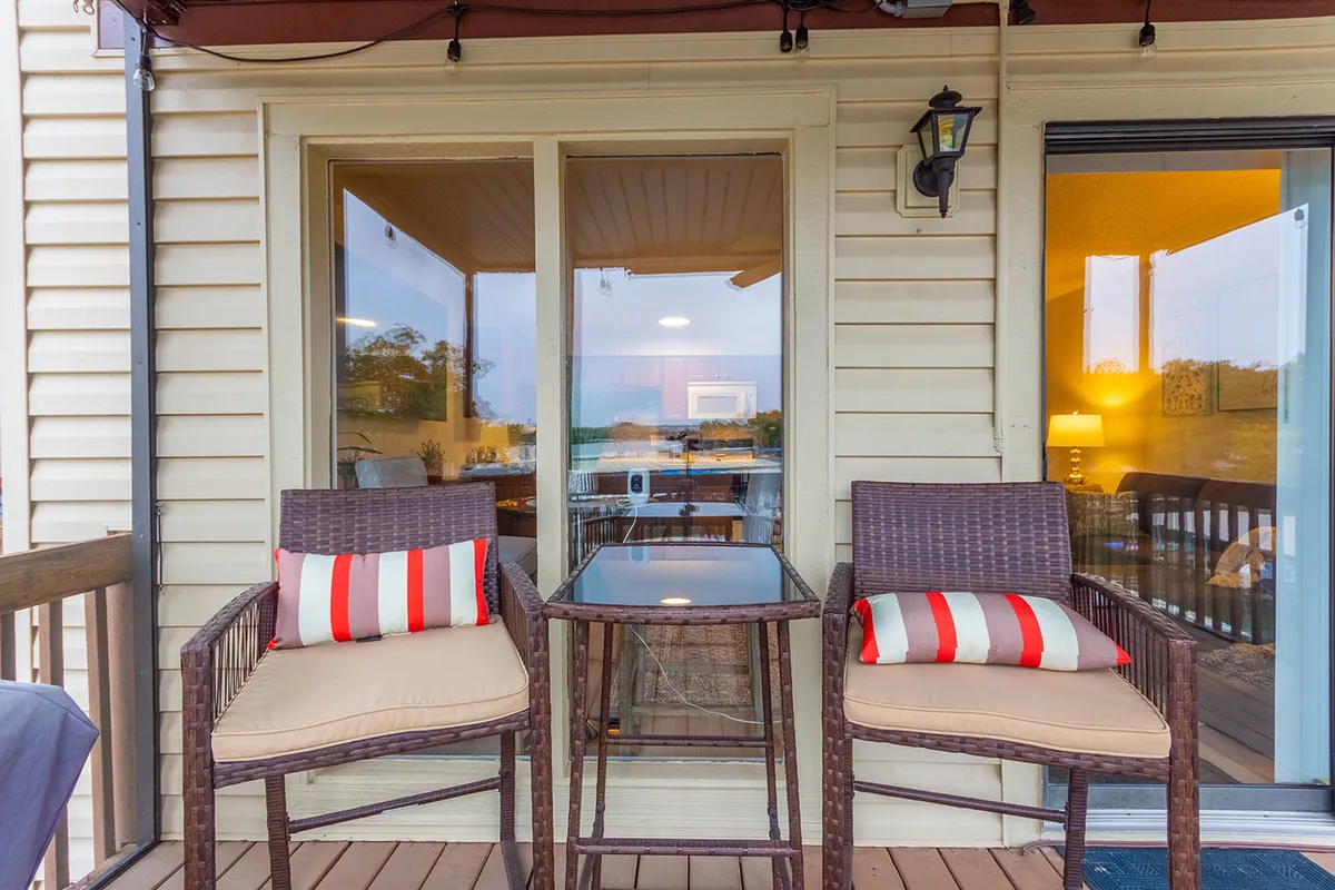 A screened-in patio with two wicker chairs, a glass table, and views of the outdoors. Large windows reveal the inviting interior beyond, and the tranquil setting is ideal for al fresco dining or enjoying coffee together while soaking in nature.