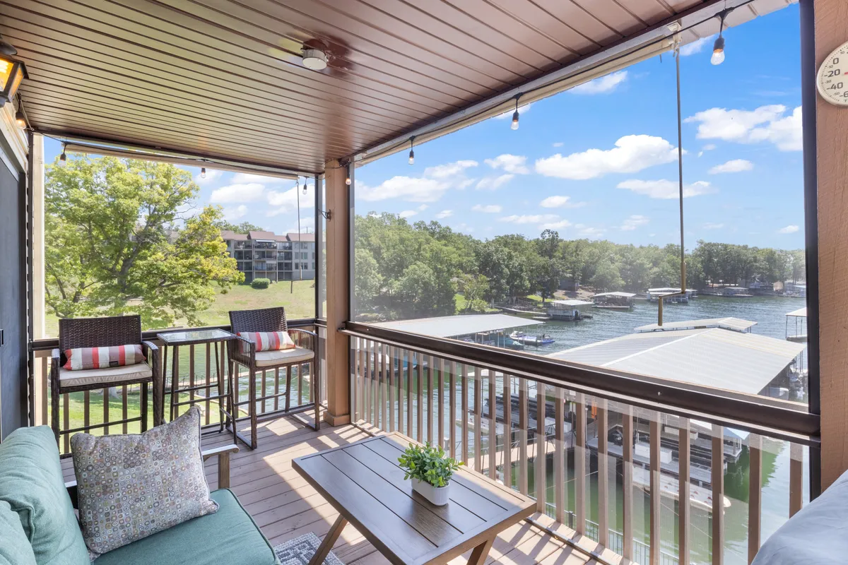 The image shows a tranquil lakefront condo exterior with a cozy seating area overlooking the water, surrounded by lush decorative plants. The space is inviting, perfect for relaxing with a book, sipping morning coffee, or enjoying lakeside sunsets. The serene lake view offers a front-row seat to passing boats and the peaceful Missouri landscape.