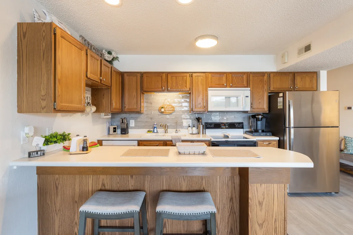 A modern kitchen featuring a central island, warm wooden cabinets, and stainless steel appliances. The space is well-lit and inviting, clearly designed for both functionality and comfort. The island offers seating for casual breakfasts or evening wine, while the appliances suggest effortless meal preparation.