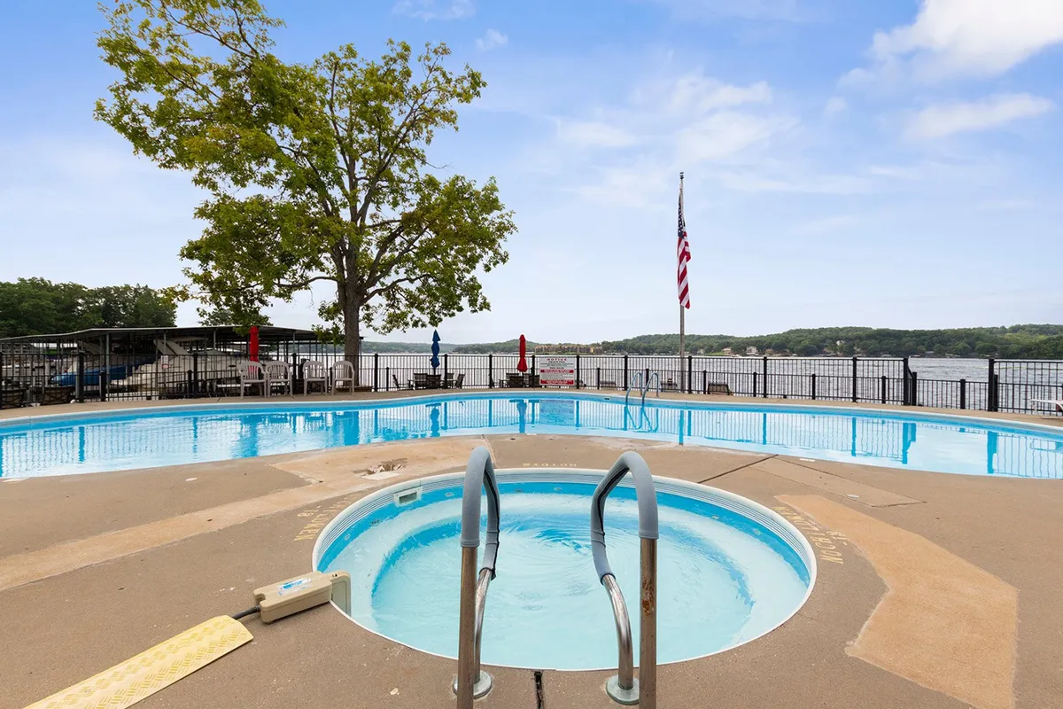 This serene exterior image depicts a sparkling pool and hot tub set against a tranquil lake backdrop, with a flag and tree adding to the peaceful vibe. The space is perfect for unwinding, socializing, or enjoying sunset views over the water.