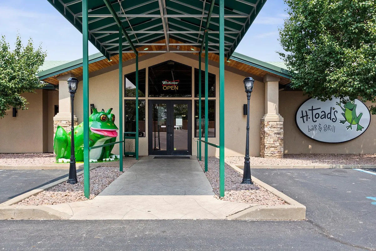 This exterior shot features the entrance of a local restaurant adorned with a large green frog statue and themed decor. The playful atmosphere suggests a lively social spot, and the signage hints at frequent live music and entertainment events popular with visitors.