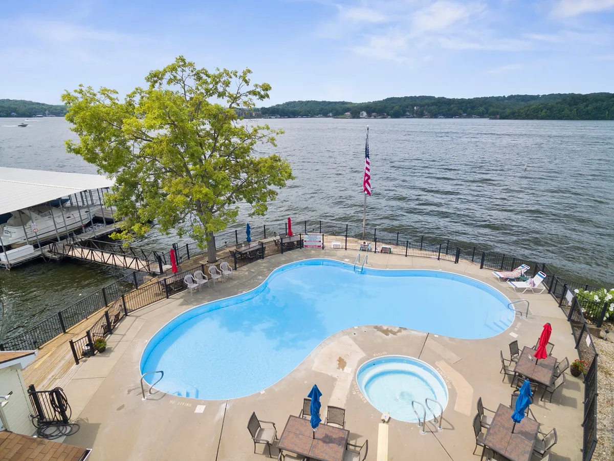 This image showcases a beautiful swimming pool and hot tub set against a picturesque lake view. Lounge chairs and a flagpole add to the inviting, resort-like atmosphere, while the water glistens in the background, evoking leisure and relaxation. The setting sun casts a tranquil glow across the scene, hinting at both daytime fun and evening serenity.