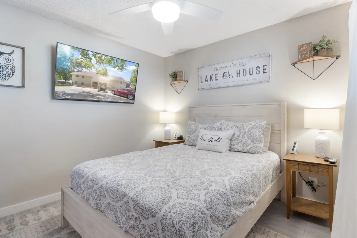 A serene bedroom featuring an intricately made bed with plush bedding, framed artwork above the headboard, nightstands with soft-glow lamps on each side, and a wall-mounted TV. Natural light filters in from a nearby window, enhancing the calming, restful atmosphere.