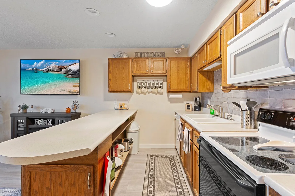 A stylish kitchen with a central island, coffee station, wall-mounted TV, and decorative signage. The space is inviting and practical, suggesting a natural flow from living to kitchen for casual dining or morning routines.