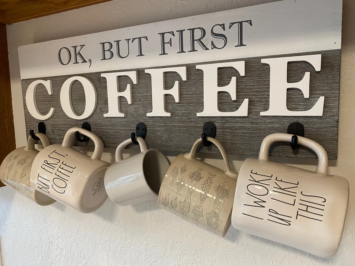 A close-up of a decorative mug rack with mugs featuring playful sayings and a wooden sign, set against the backdrop of a cozy kitchen. The arrangement evokes a sense of personalized comfort and shared morning coffee routines.