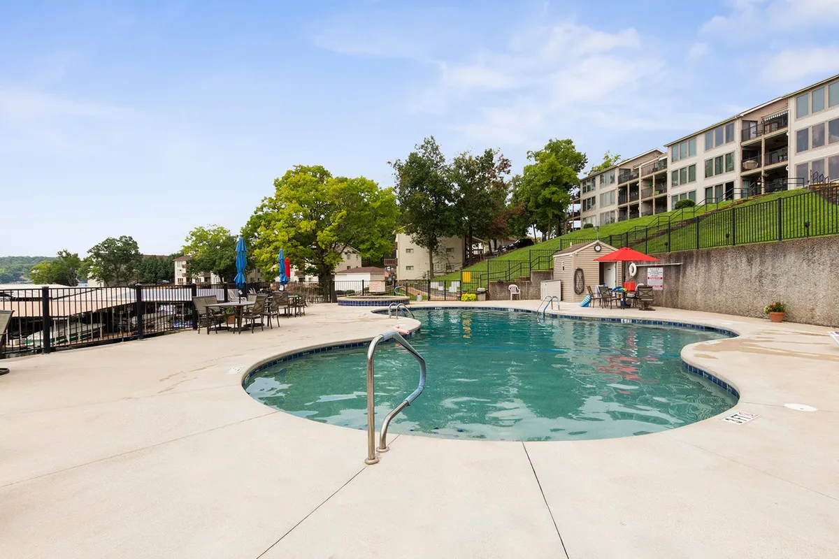A sparkling swimming pool takes center stage, surrounded by lounge chairs and large umbrellas that invite guests to relax and soak up the sun. Lush greenery borders the pool area, creating a tranquil and resort-like atmosphere. The environment exudes a sense of relaxation and community, hinting at socializing, sunbathing, and enjoying lazy afternoons by the water.